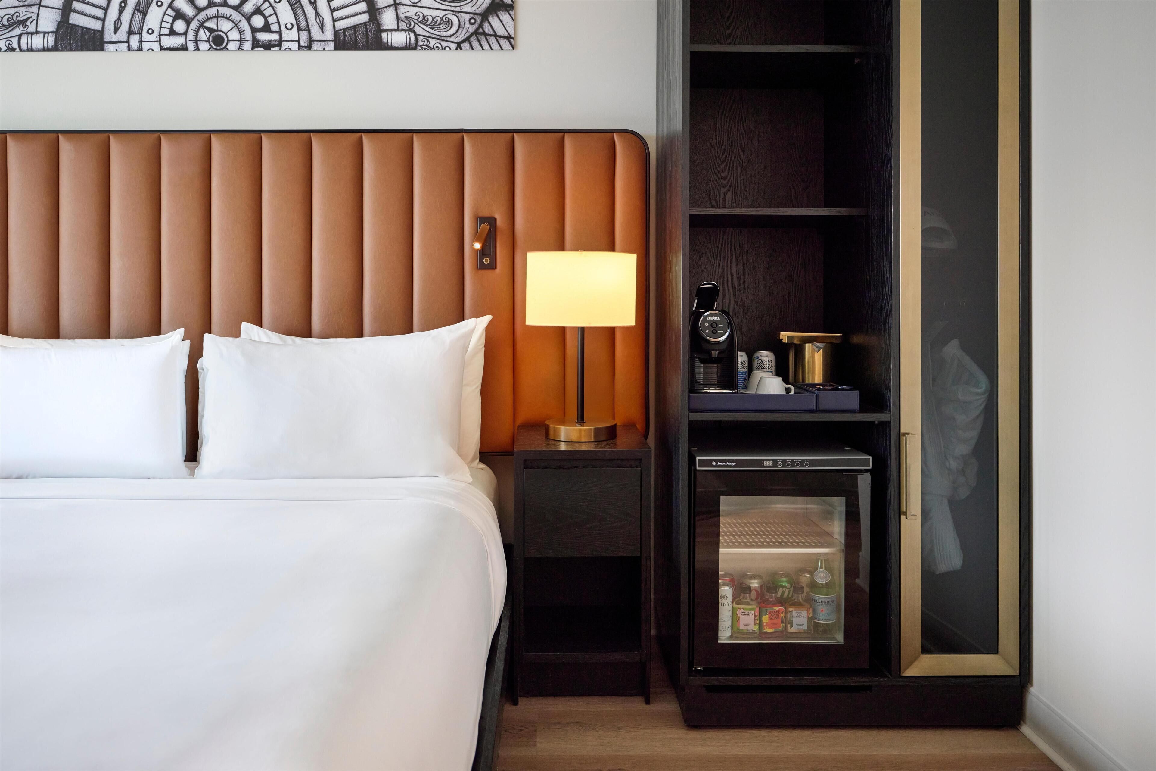 Habitación de hotel con cama, mesita de noche con lámpara, minibar y una obra de arte colgada en la pared.