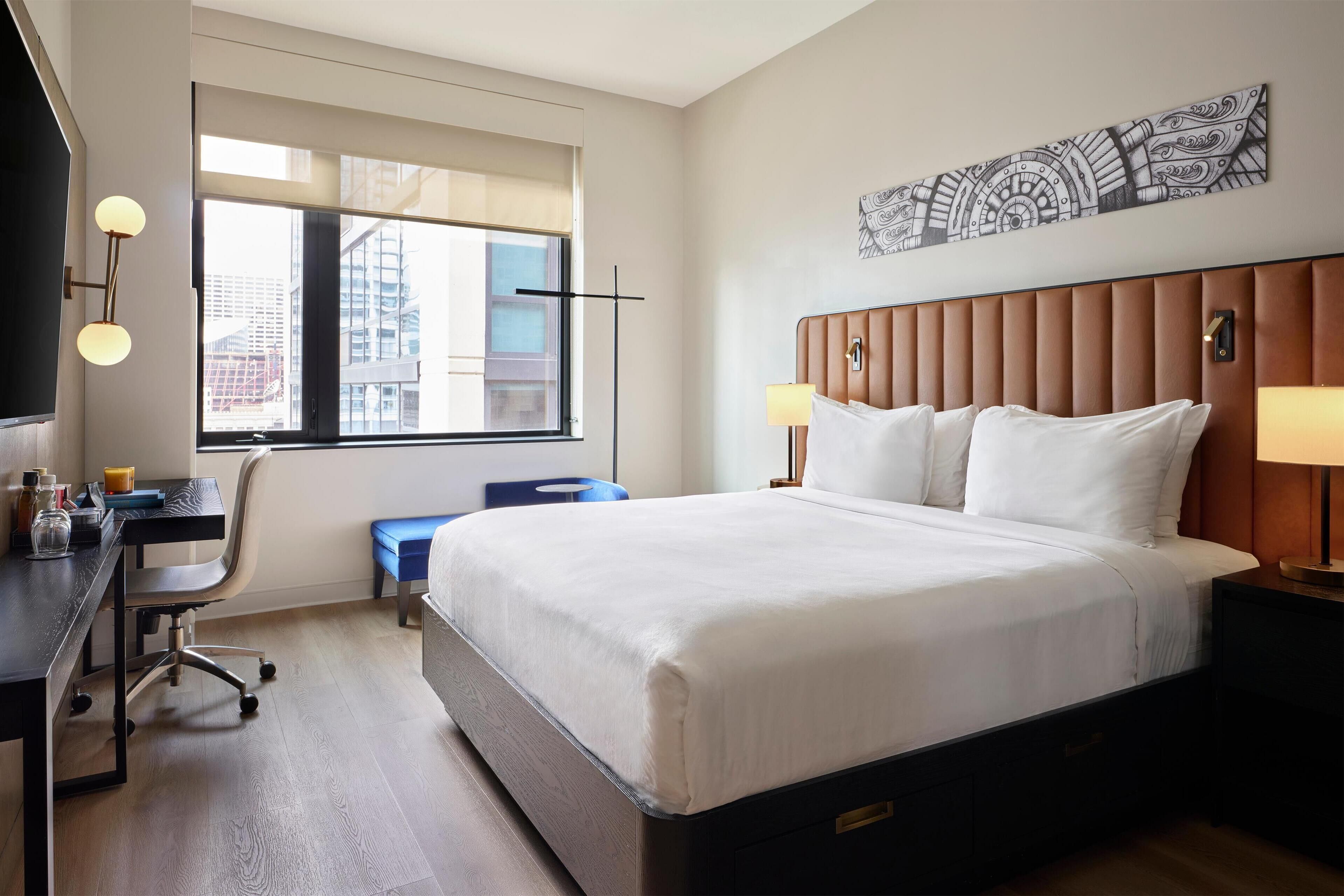 A hotel room with a large bed, a desk with a chair, a window with a view of buildings, and a wall-mounted artwork.