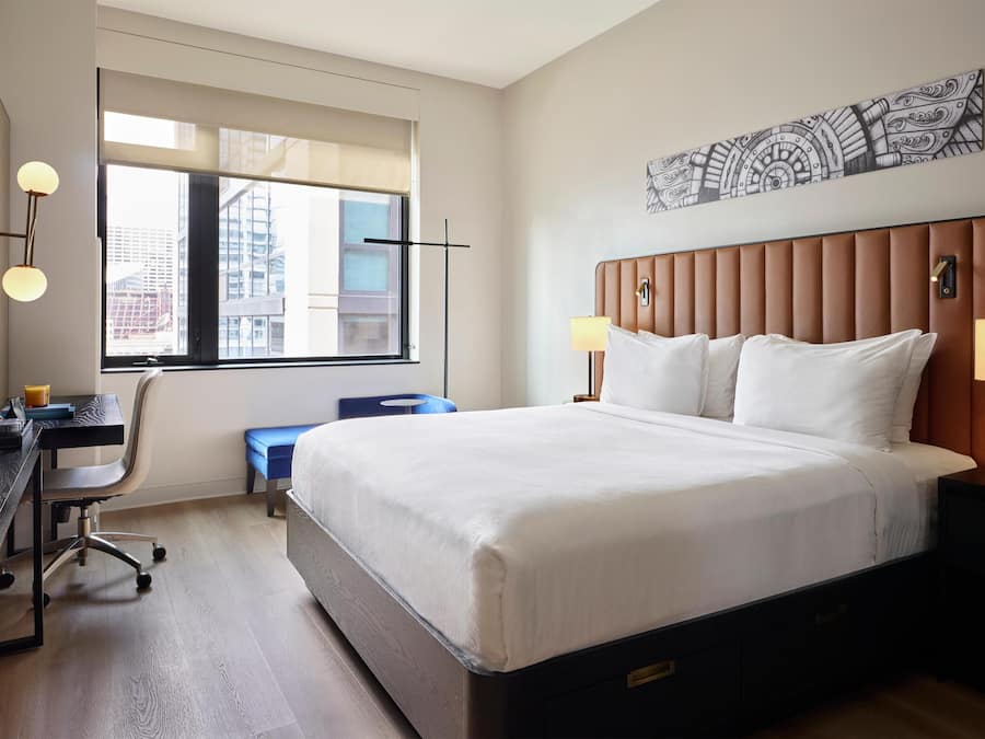 A hotel room with a large bed, a desk with a chair, a window with a view of buildings, and a wall-mounted artwork.