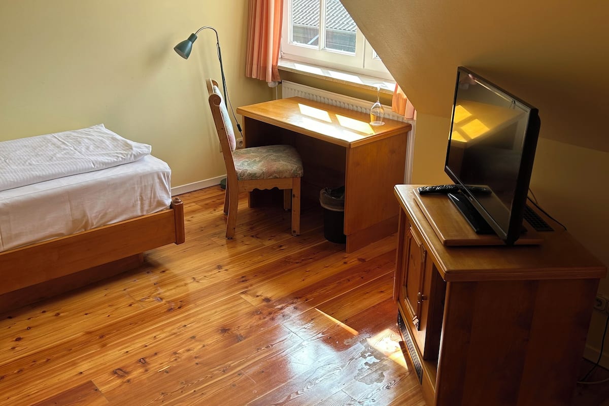 A small, cozy room with a wooden floor, a bed, a desk with a chair, a television, and a window with curtains.