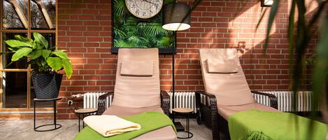 A sunlit outdoor seating area with two lounge chairs, a potted plant, a wall clock, and a brick wall.