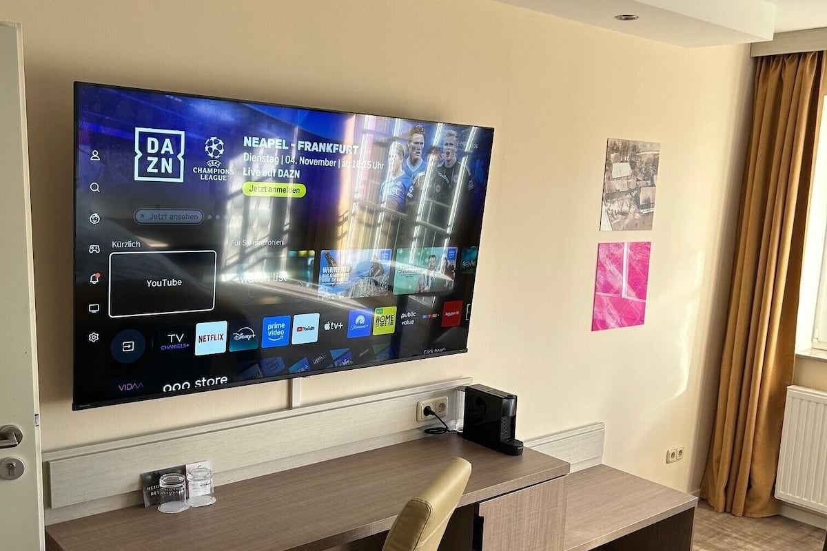 A modern hotel room with a large flat-screen TV mounted on the wall, a wooden desk with a chair, and a radiator.