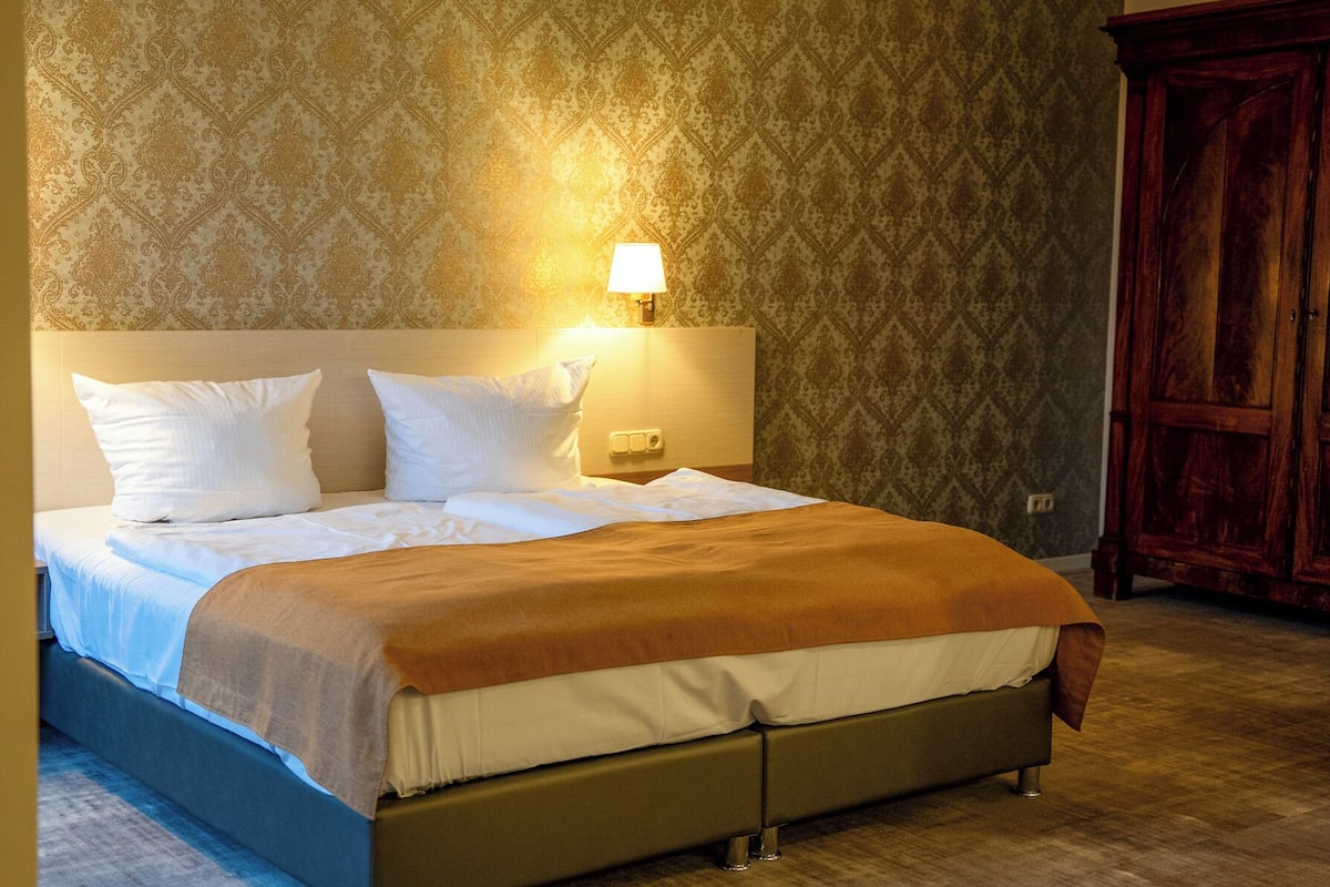 A neatly made bed with white linens and a brown throw blanket, a wall-mounted lamp, and a wooden wardrobe.