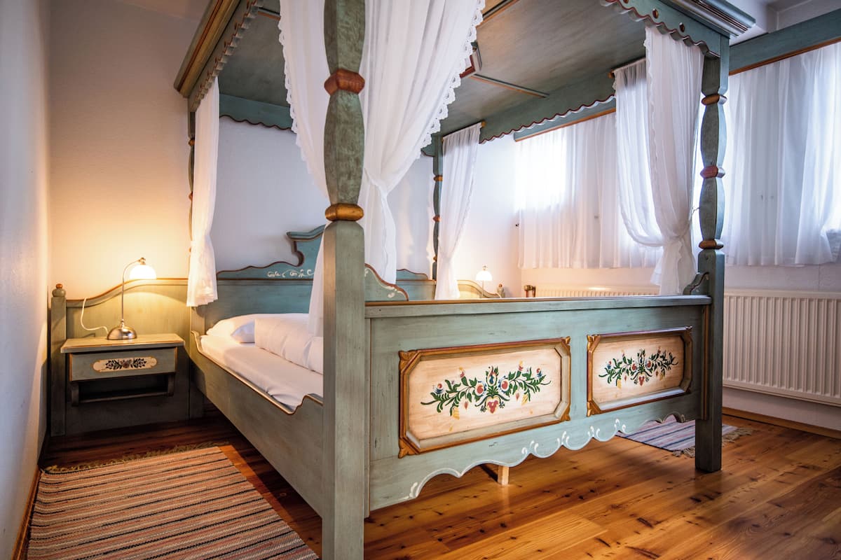 A four-poster bed with a wooden frame and floral paneling, a bedside table with a lamp, and a window with sheer curtains.