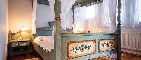 A four-poster bed with a wooden frame and floral paneling, a bedside table with a lamp, and a window with sheer curtains.