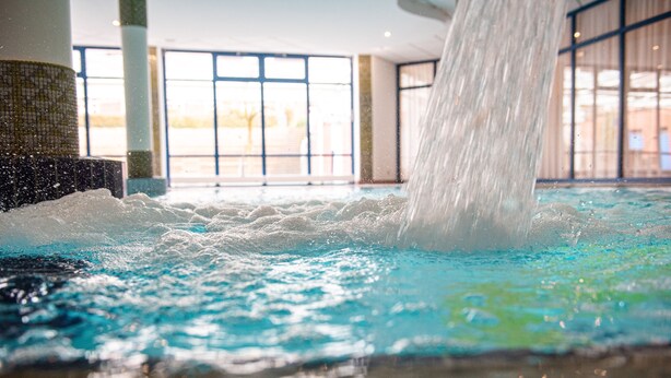 Un élément décoratif aquatique avec une cascade, situé dans un vaste espace intérieur abritant une piscine.