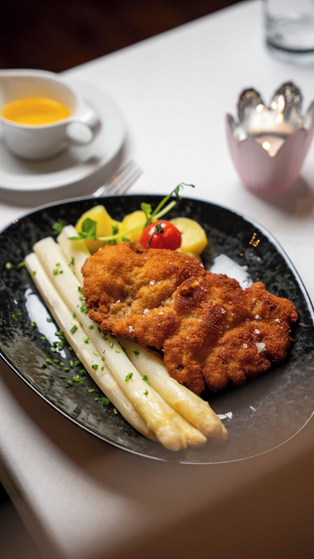 A plate with breaded cutlet, asparagus, lemon, and cherry tomato garnished with herbs.