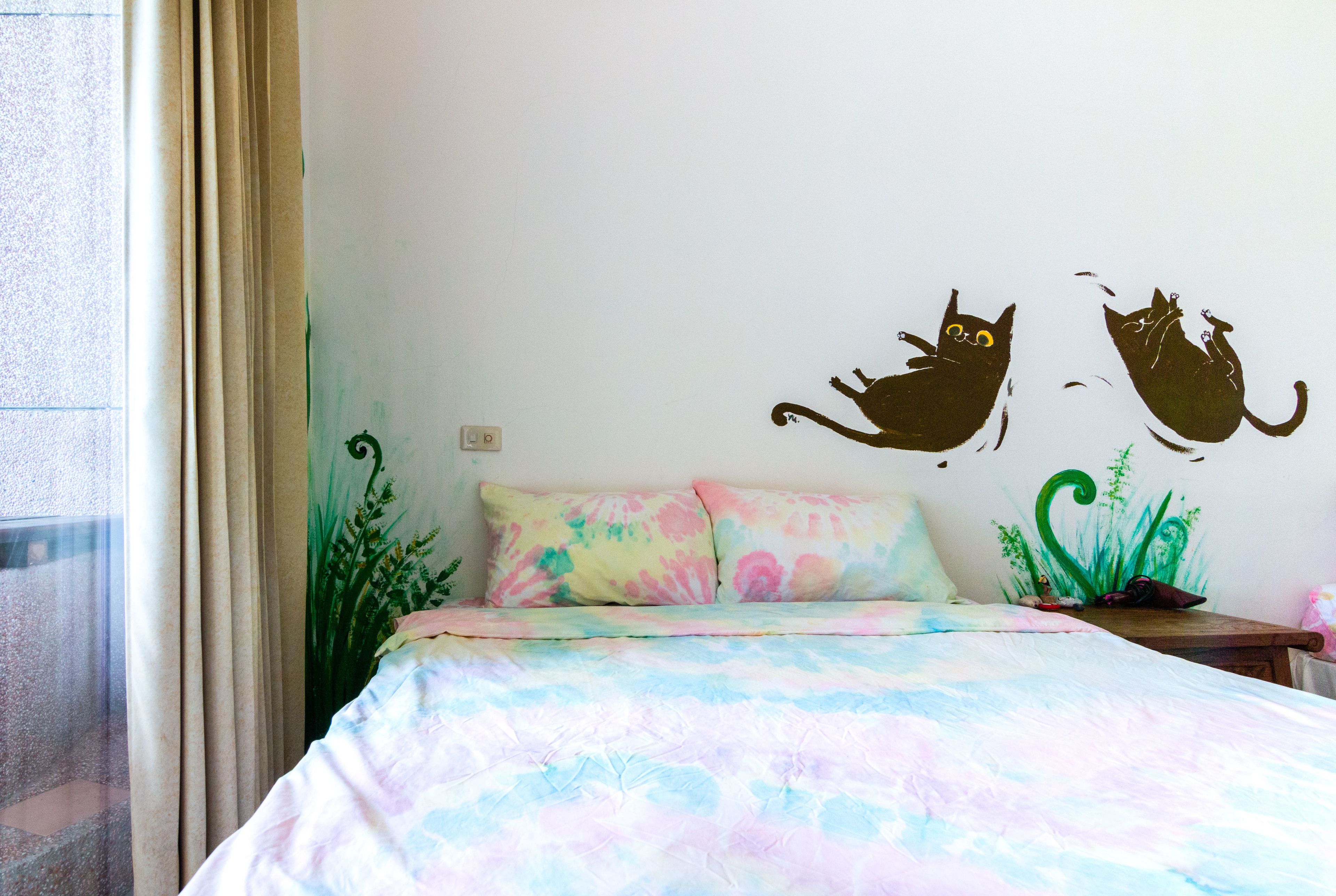 A bedroom with a bed, two pillows, a nightstand, a plant, and wall decals of cats.