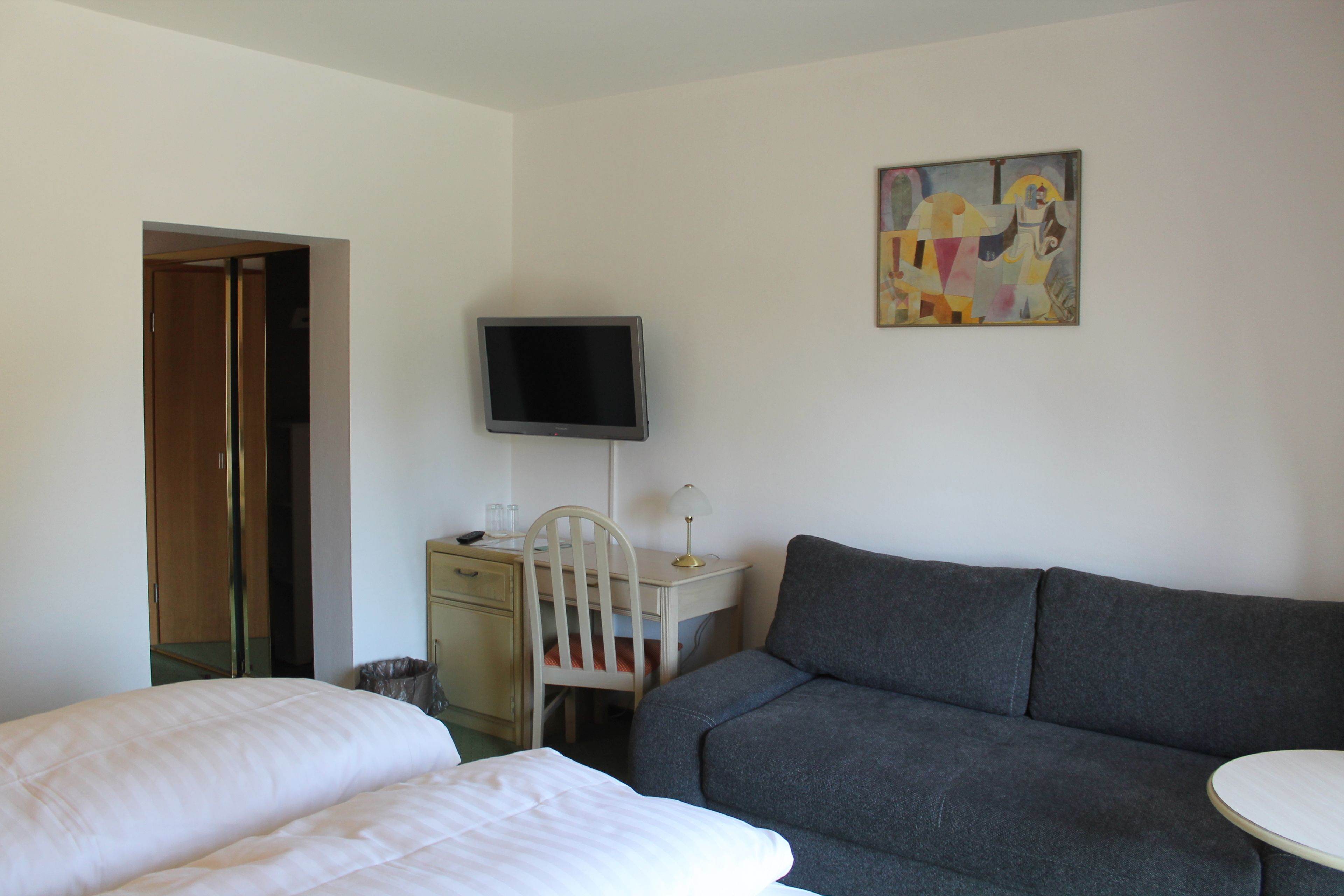 A hotel room with a bed, a grey sofa, a desk with a computer and a TV, and a painting on the wall.