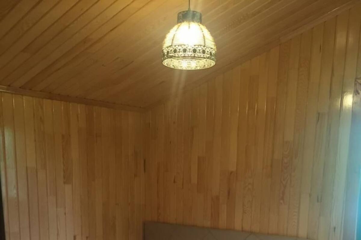 Ein Zimmer mit Holzwandverkleidung, einem Bett mit weißer Matratze und einer pinkfarbenen, strukturierten Decke sowie einer hängenden Leuchte.