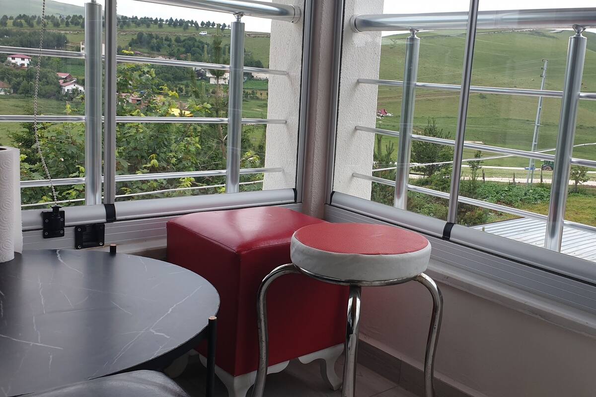 Ein Zimmer mit einem großen Fenster, das Blick auf eine grüne Landschaft und eine Straße bietet. Es gibt einen Tisch mit einem Stuhl und einem roten Hocker.