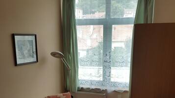 Una habitación pequeña y ordenada con una cama, una silla, un escritorio, un televisor y una ventana con vistas a edificios.