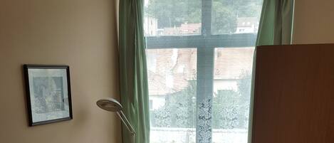 A small, neatly arranged room with a bed, a chair, a desk, a television, and a window with a view of buildings outside.