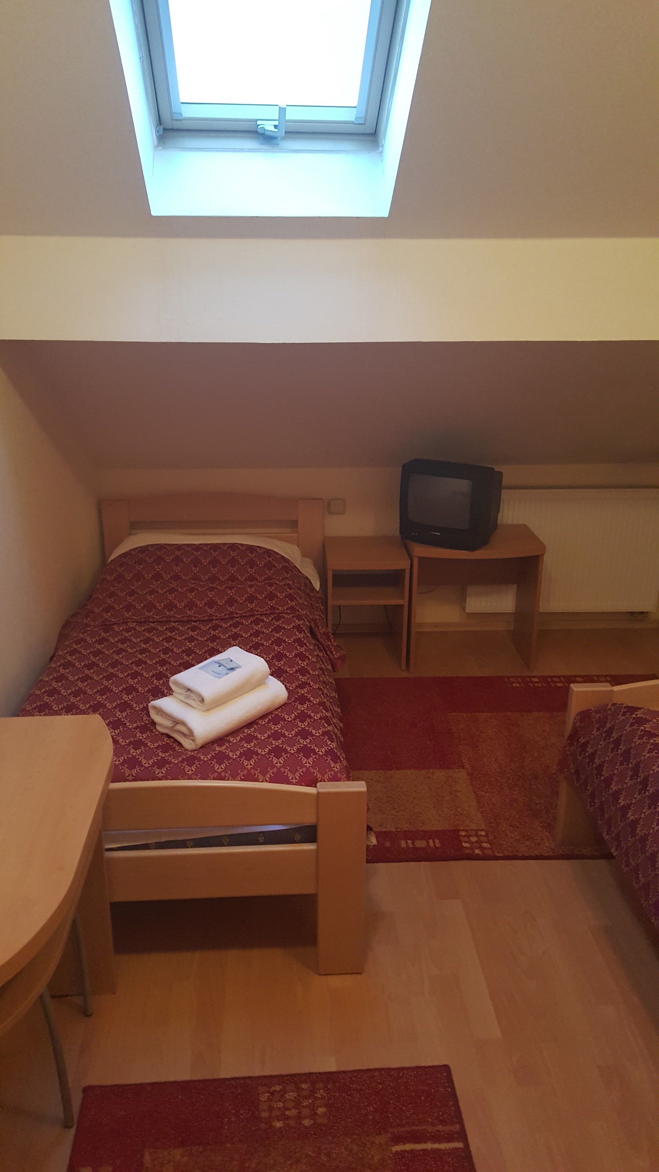 Un dormitorio con tragaluz, una cama con colcha estampada roja, una mesita de noche de madera con televisor, y una alfombra roja en el suelo.
