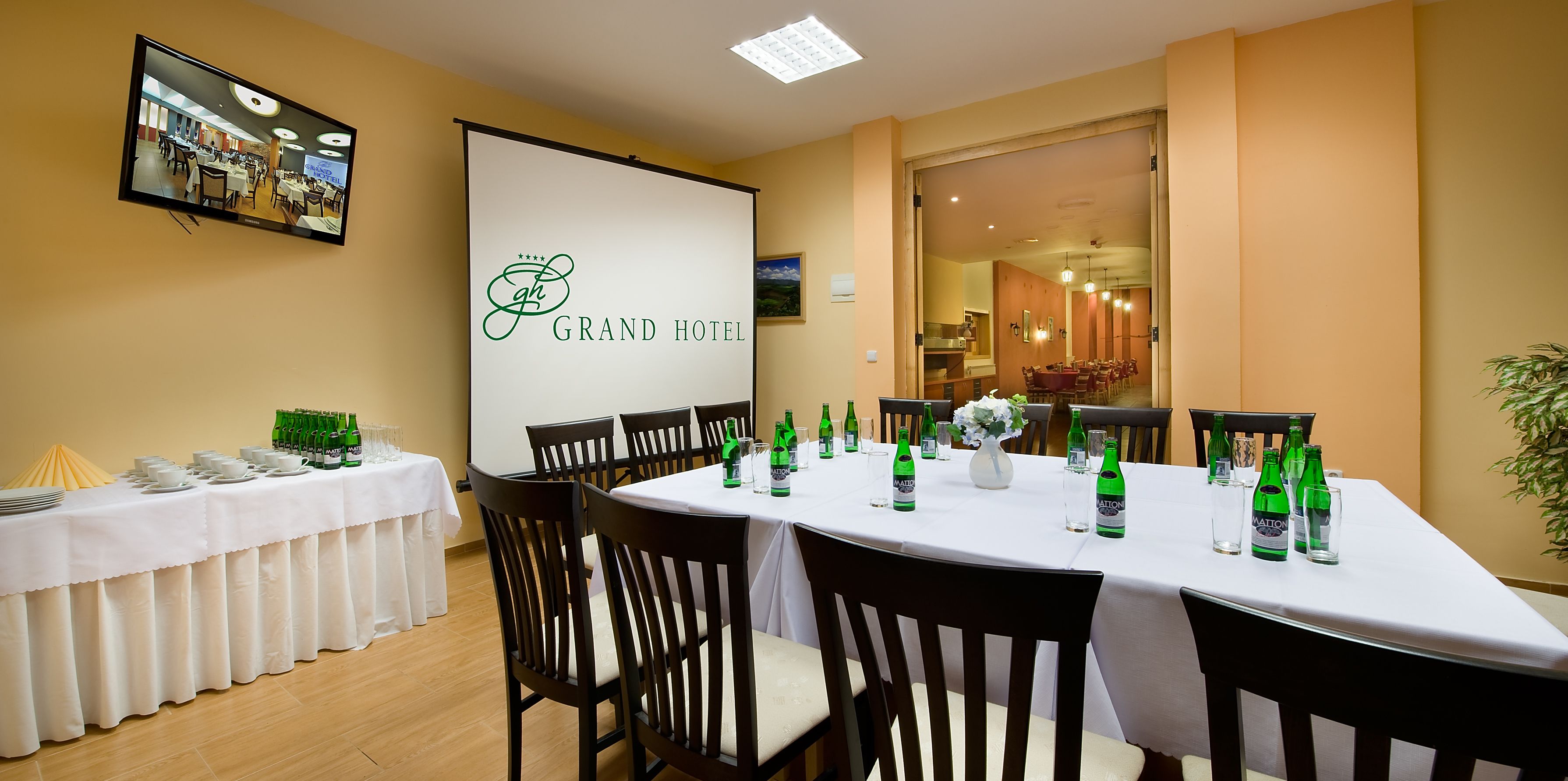 Una sala conferenze con un tavolo bianco, sedie e uno schermo proiettore che mostra il logo del Grand Hotel.