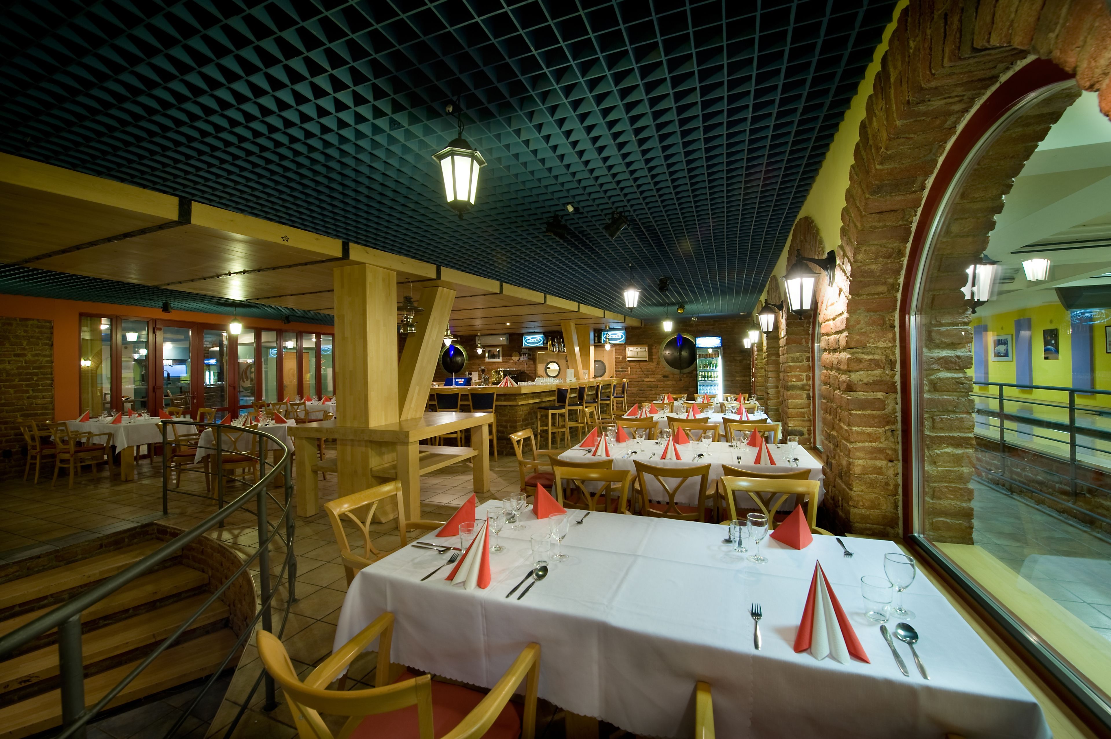 A restaurant with multiple dining tables set with white tablecloths and red napkins, brick walls, and a view of a train through a large arched window.