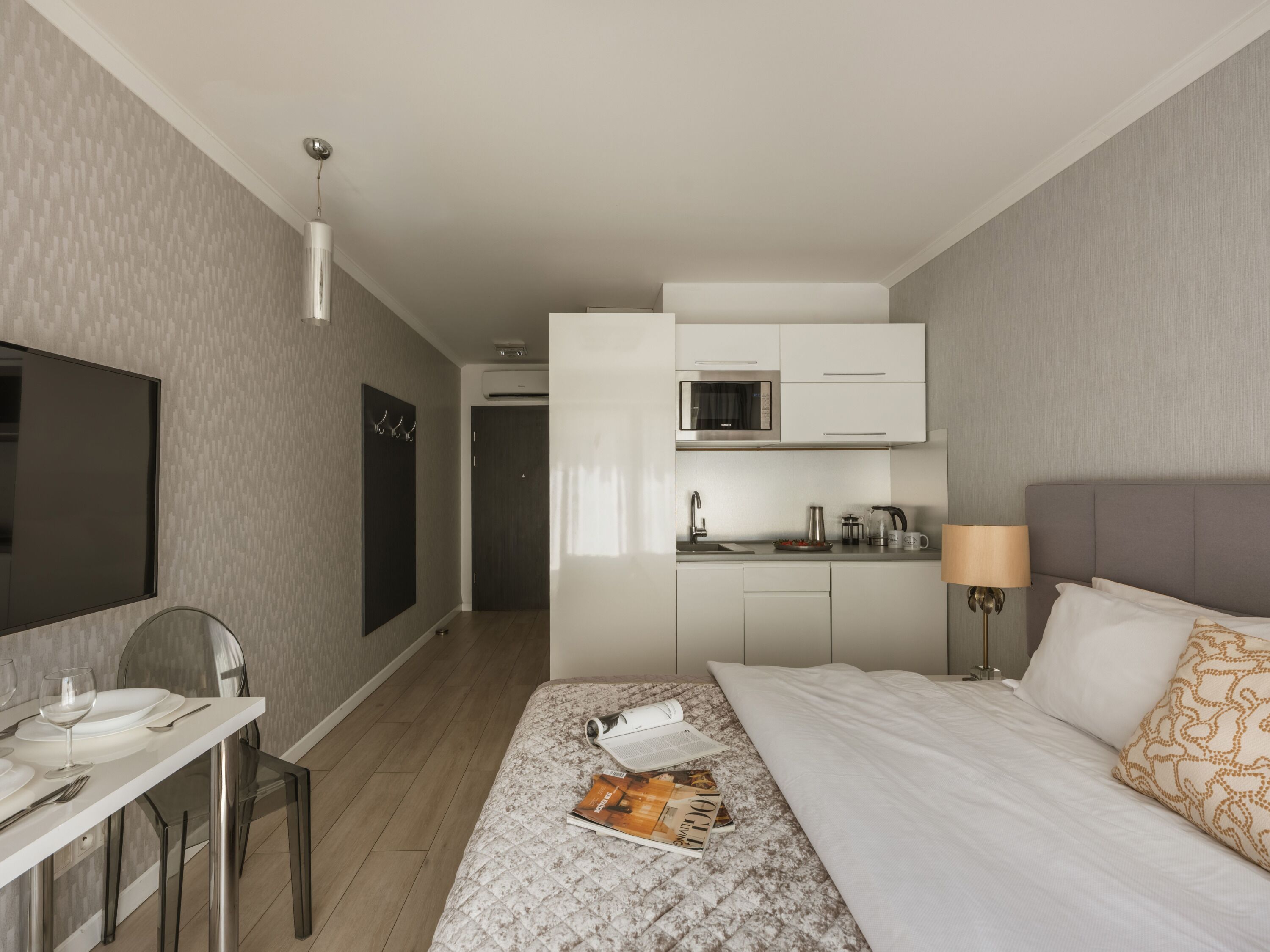 A hotel room with a bed, a desk with a chair, a kitchenette, and a TV.