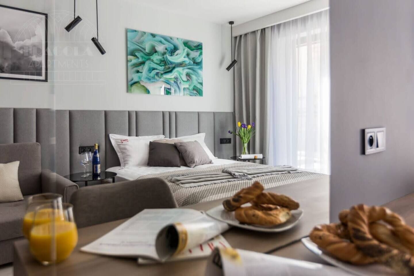 A modern hotel room with a bed, a sofa, a dining table with breakfast, and a large abstract painting.