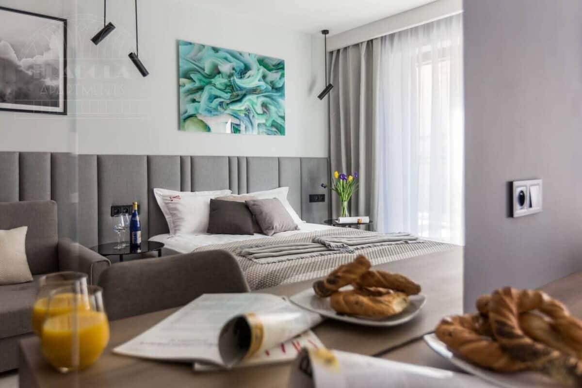 A modern hotel room with a bed, a sofa, a dining table with breakfast, and a large abstract painting.
