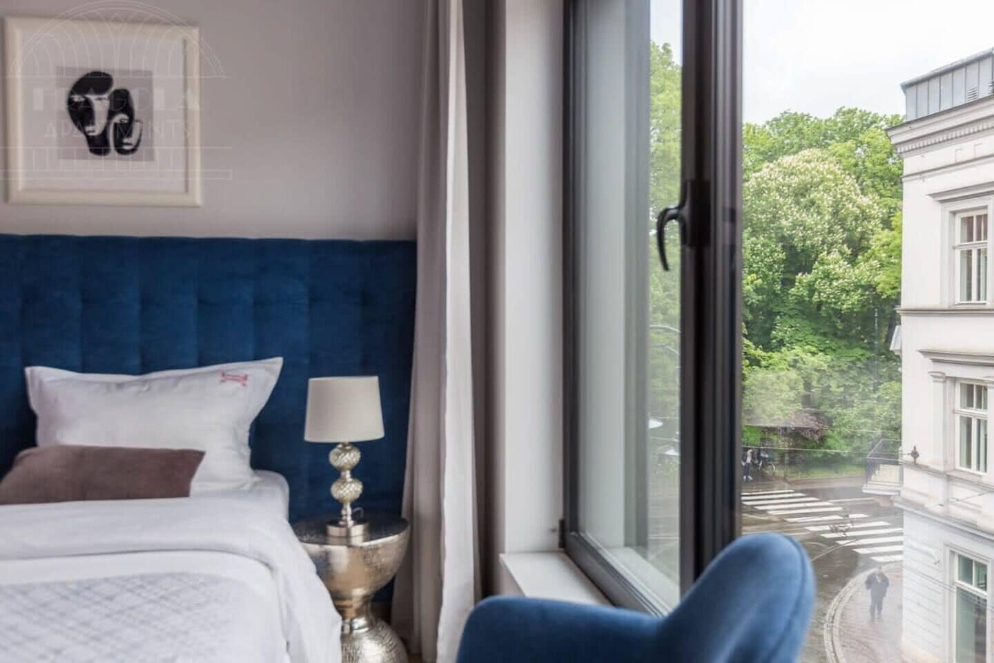 A hotel room with a large window, a bed with white bedding, a blue headboard, and a framed artwork on the wall.