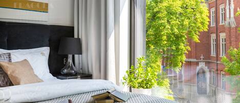 A bedroom with a bed, a nightstand, a lamp, a window with a view of a red brick building and a tree.
