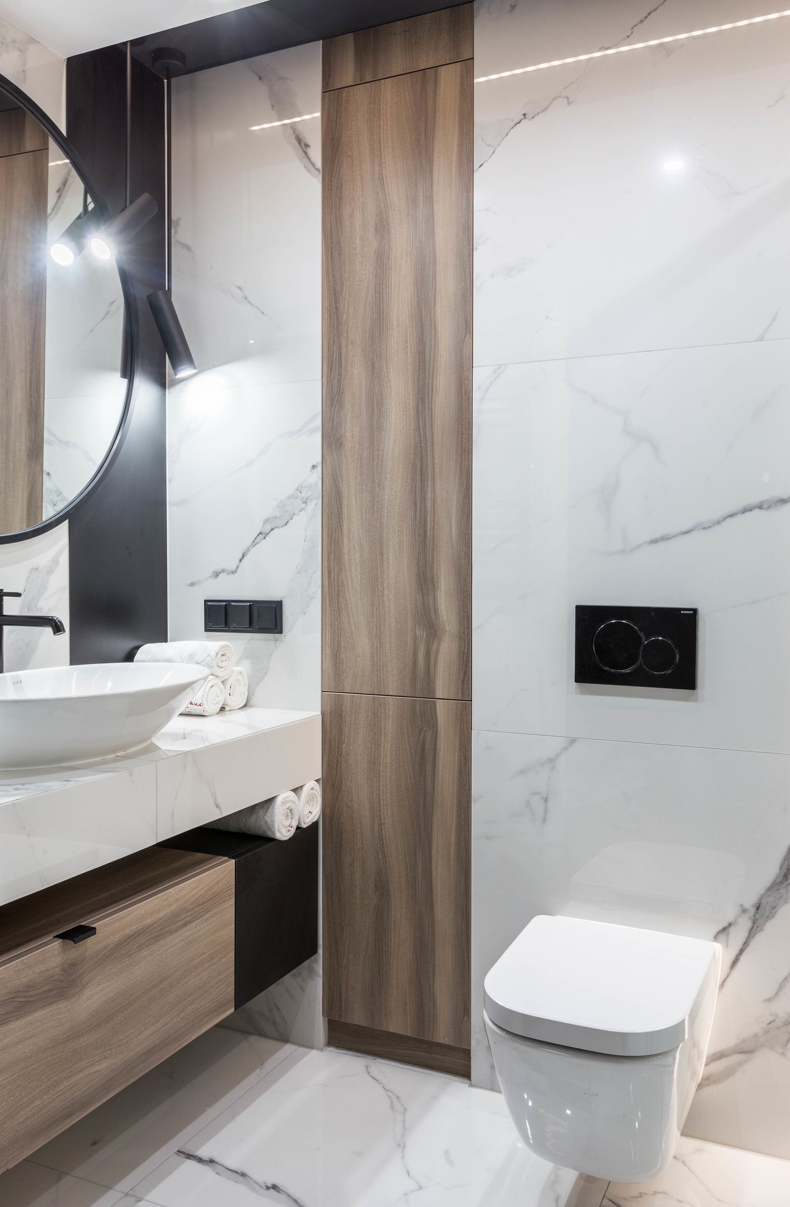 A modern bathroom with a white sink, wooden vanity, a round mirror, and a toilet.
