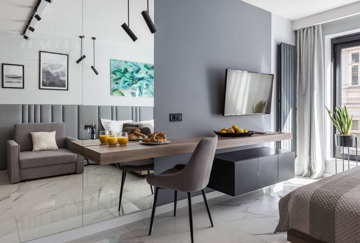 A modern hotel room with a dining area, a sofa, a TV, and a large window.