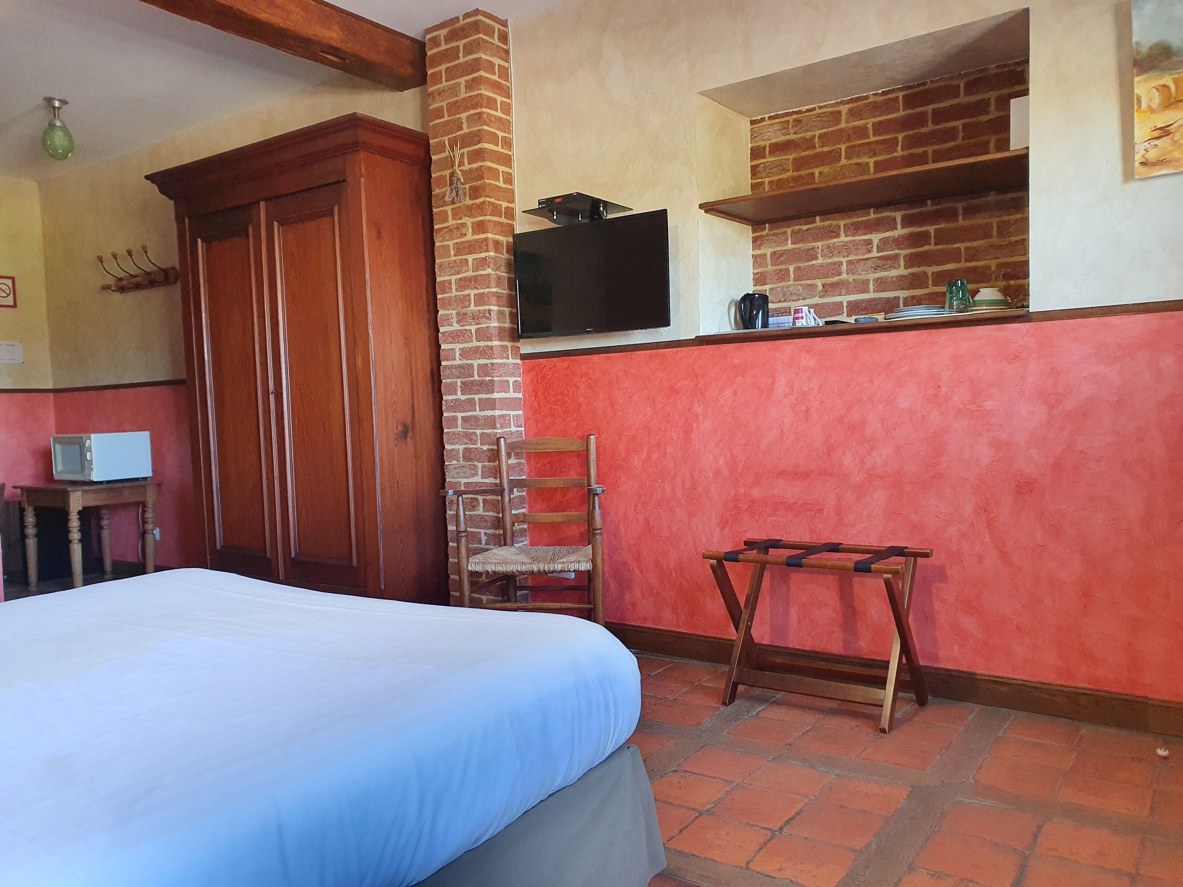 A room with a bed, a wooden chair, a small table with a microwave, a brick wall, and a television mounted on the wall.