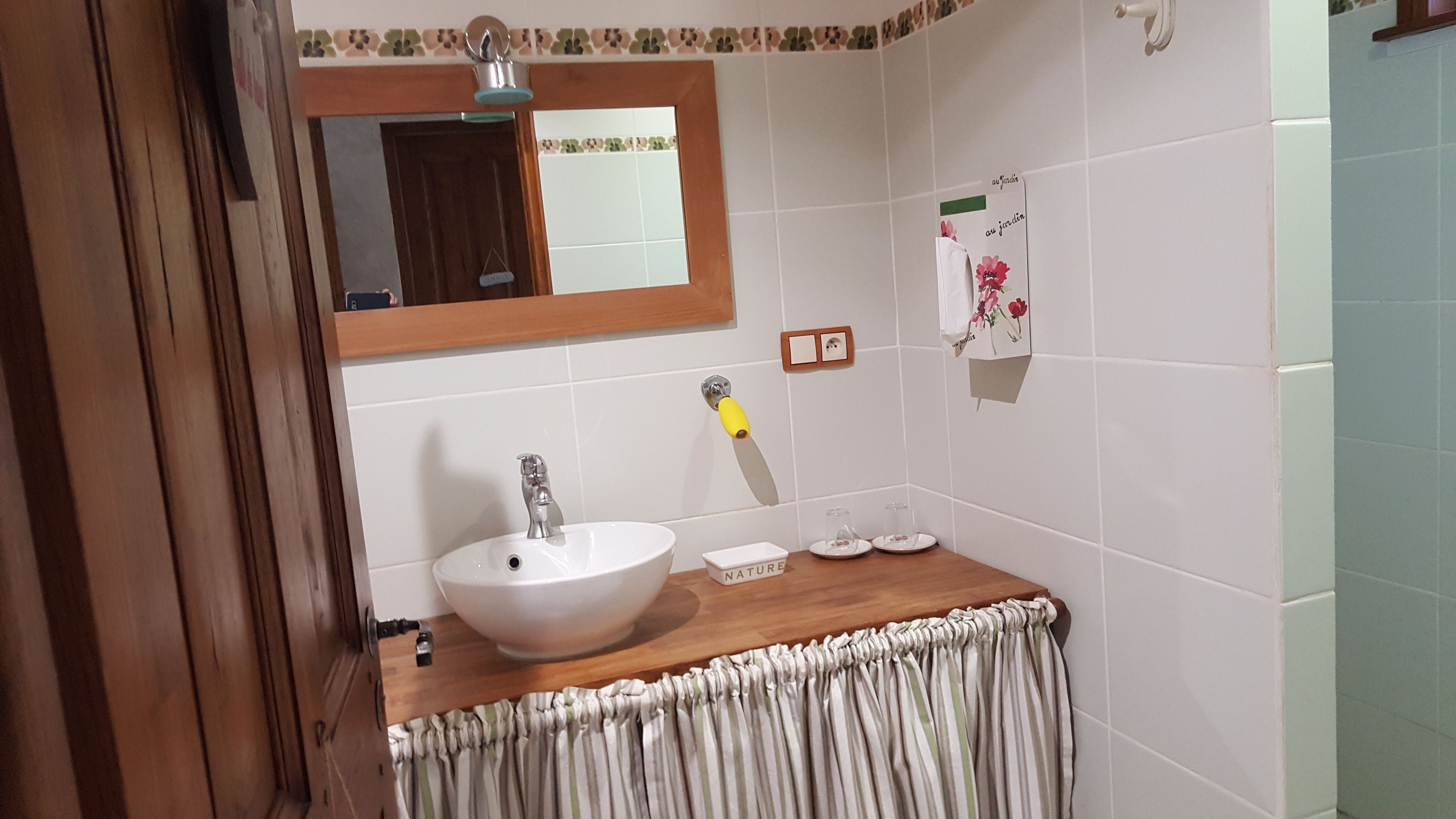 A bathroom with a white sink, wooden countertop, and a mirror above it. There is a towel rack on the wall and a small shelf with a glass container.