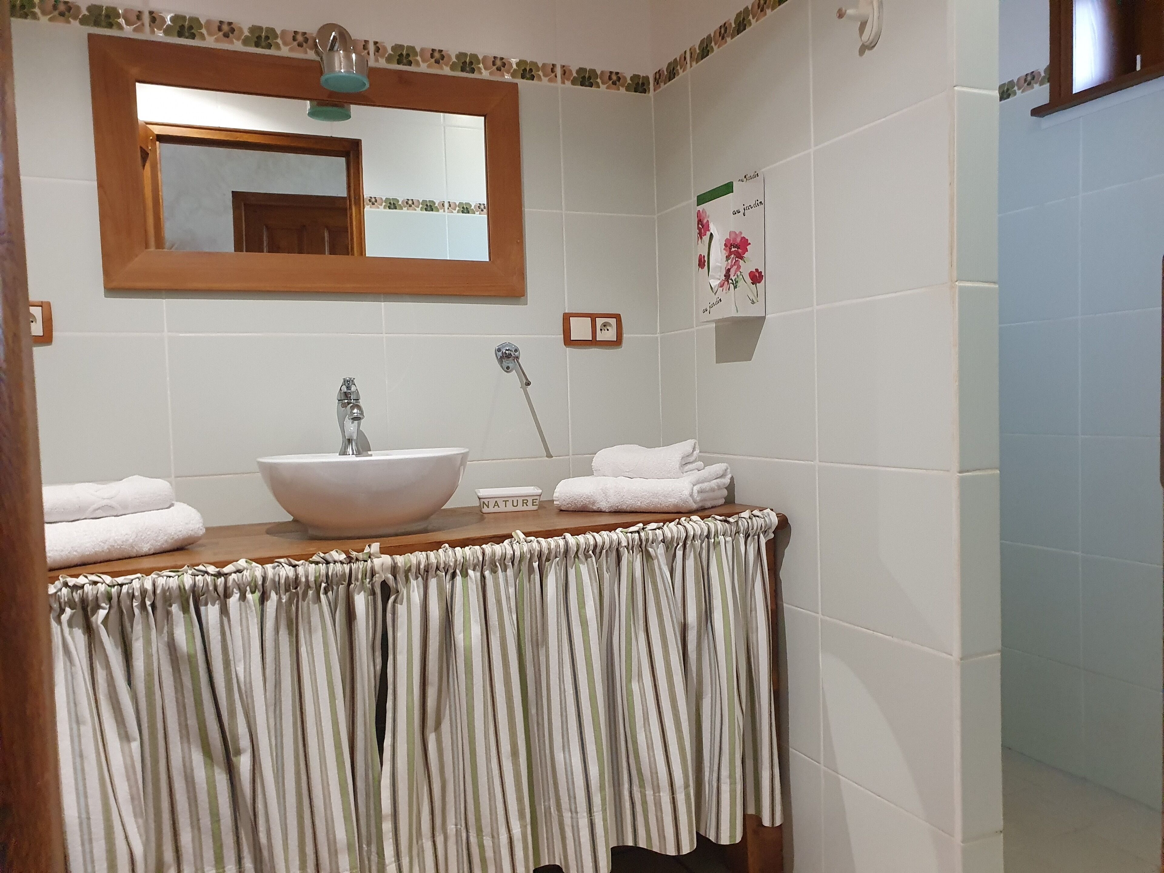 A bathroom with a wooden vanity, a white sink, and a mirror above it. There are towels stacked on the vanity and a small sign on the wall.