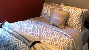 Ein ordentlich gemachtes Bett mit gemusterten Bettwäsche, ein Kissen mit dem Schriftzug 'fantasy' und zwei gerahmte Kunstwerke an der Wand.