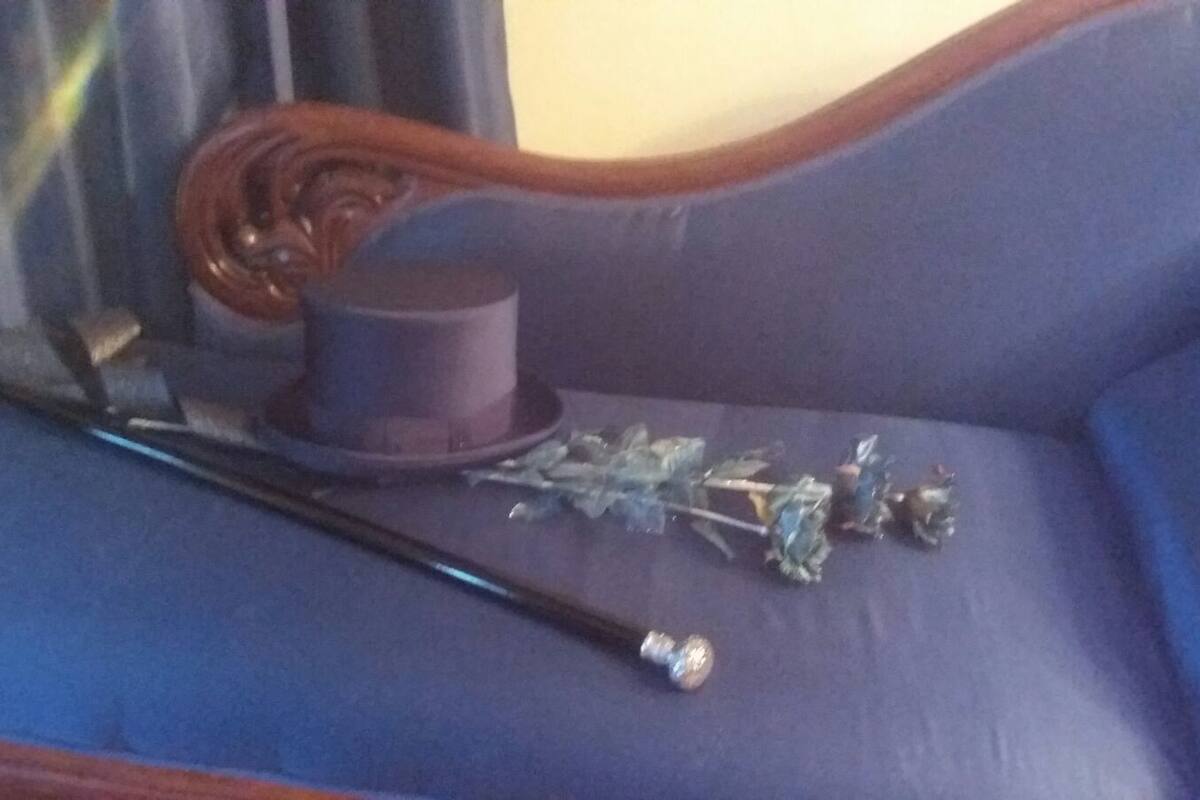 Un canapé bleu avec un accoudoir en bois, une canne noire, un haut-de-forme violet et un bouquet de fleurs.