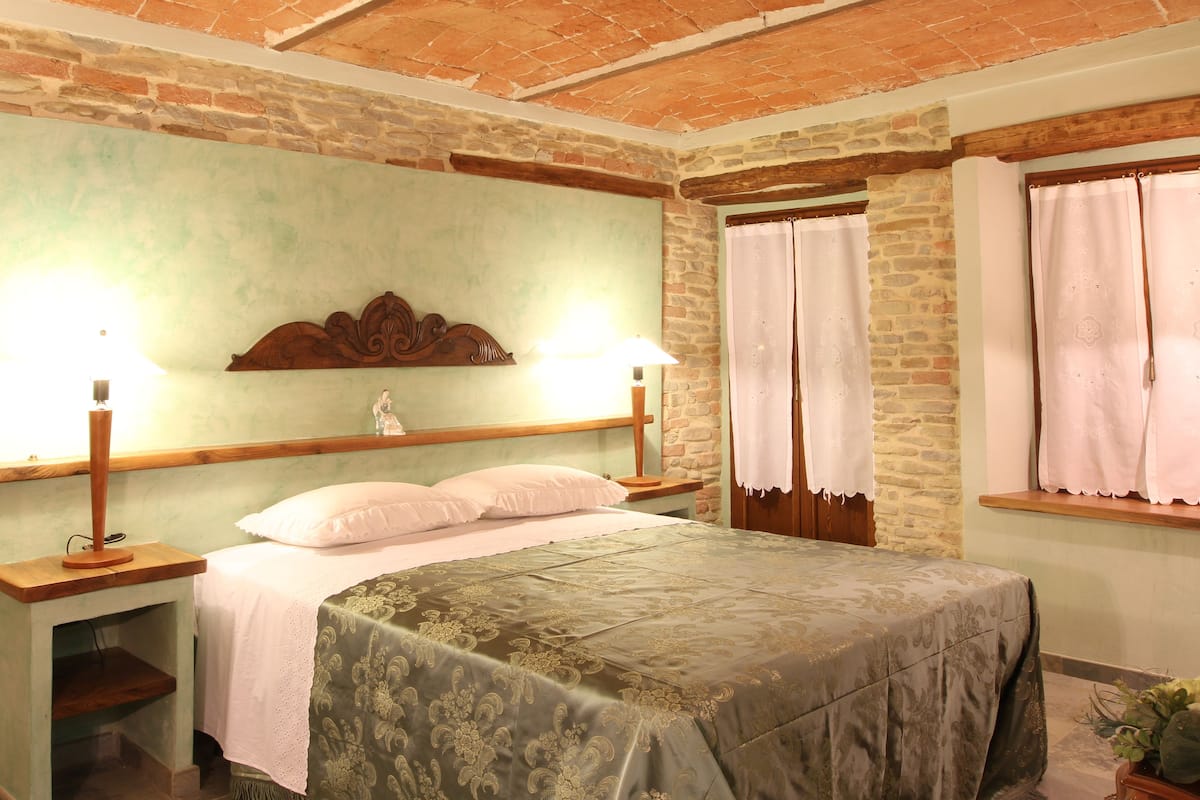 Une chambre avec un lit, une tête de lit en bois, deux tables de chevet, une commode, une lampe, une fenêtre avec des rideaux et un mur en briques.