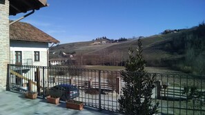 A balcony with a view of a vineyard, a parked car, and a house in the background. - Agriturismo Ercolana (Neviglie)