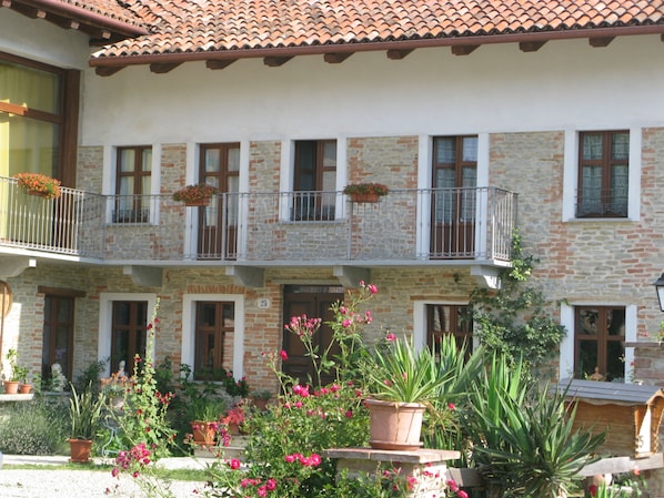 A two-story building with a stone facade, multiple balconies with flower boxes, and a garden area with potted plants and flowers. - Agriturismo Ercolana (Neviglie)