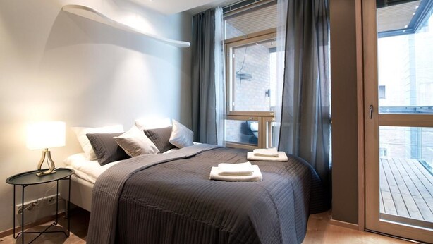 A modern hotel room with a large bed, a bedside table with a lamp, a window with curtains, and a view of the outside balcony. - 2ndhomes Kamppi Center Apartment (Helsinki)