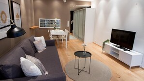 A modern living room with a grey sofa, a round coffee table, a TV on a white stand, and a dining area with a white table and chairs. - 2ndhomes Kamppi Center Apartment (Helsinki)