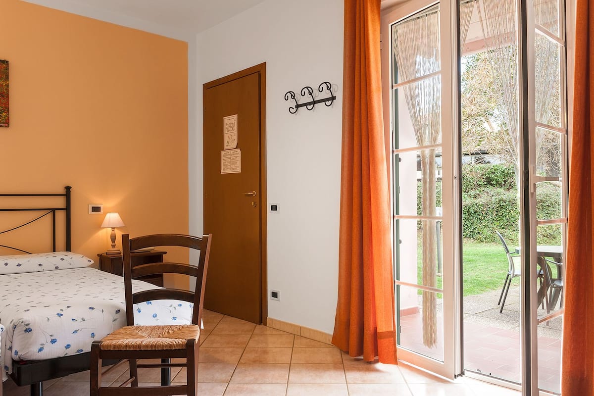 A bedroom with a bed, a chair, a nightstand, a lamp, a door with a sign, and a view of the outdoors through a glass door.