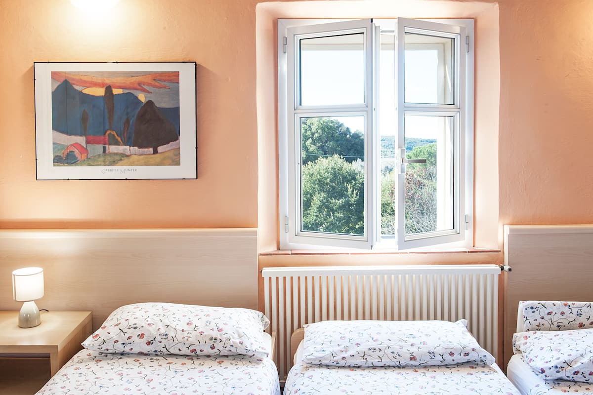 A bedroom with two beds, a window with a view of greenery, a painting on the wall, and a bedside lamp.