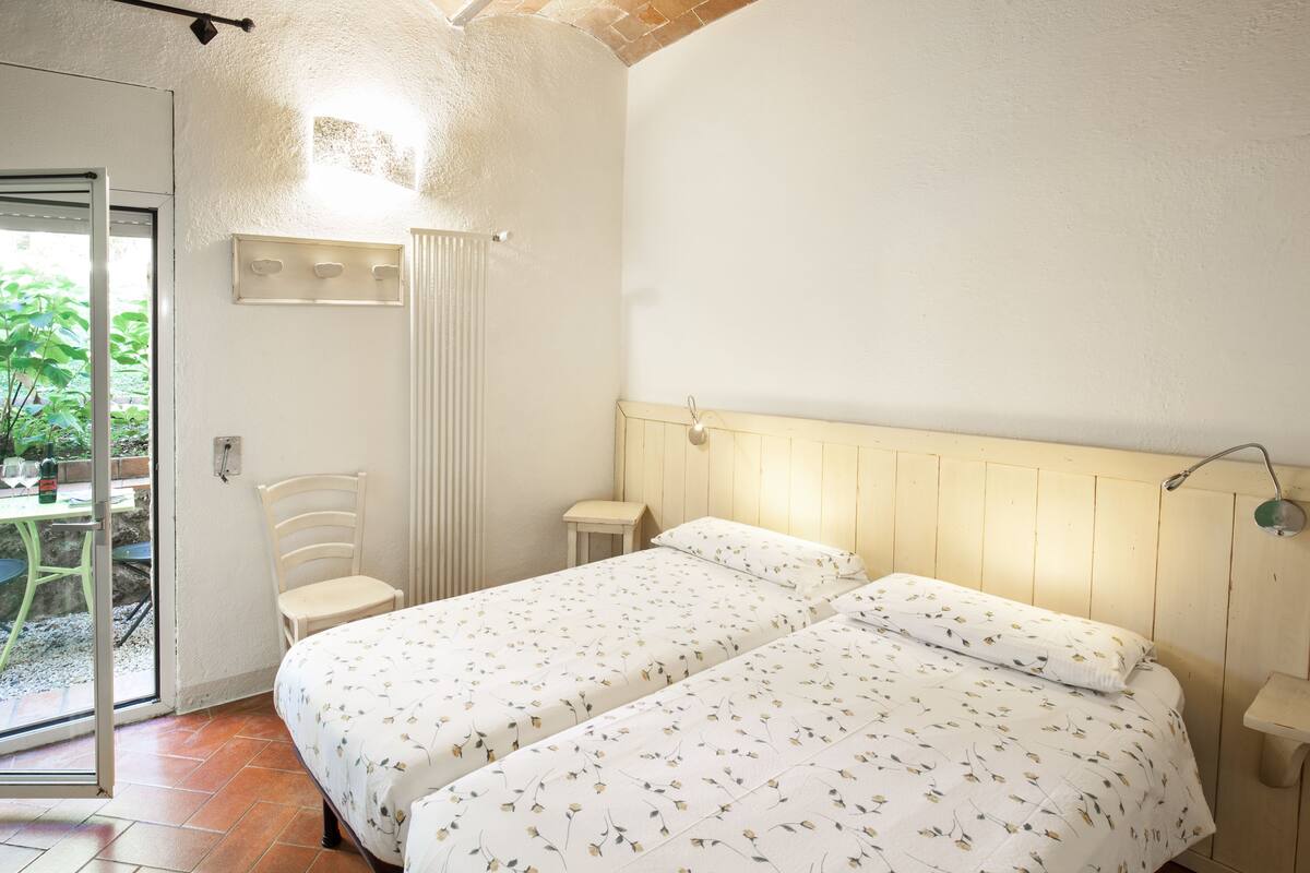 A bedroom with two beds, a chair, a table, and a window with a view of greenery.
