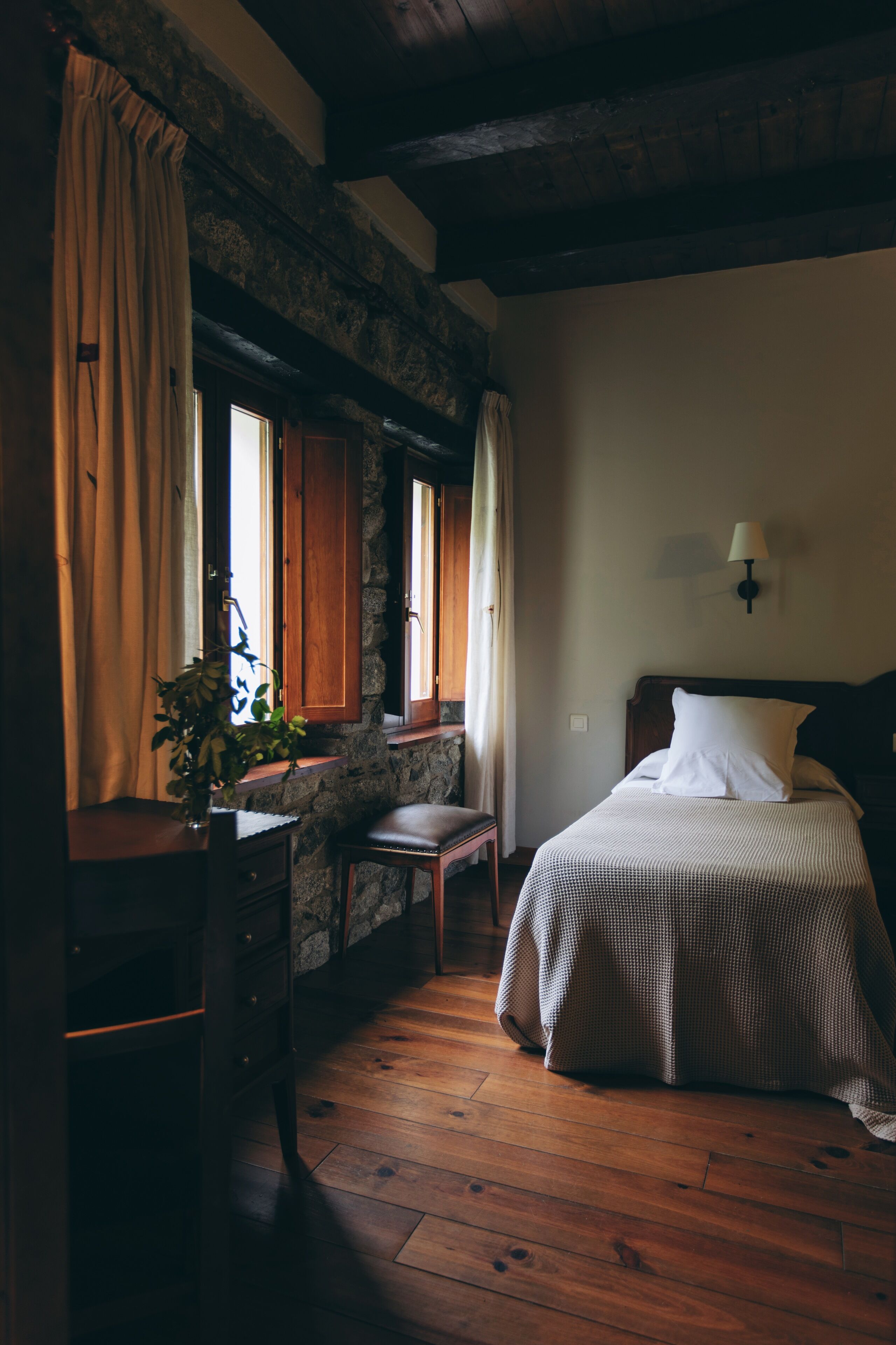 A room with a wooden floor, a bed with white bedding, a nightstand, a desk with a plant, and a stone wall.