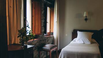 A room with a wooden floor, a bed with white bedding, a nightstand, a desk with a plant, and a stone wall.