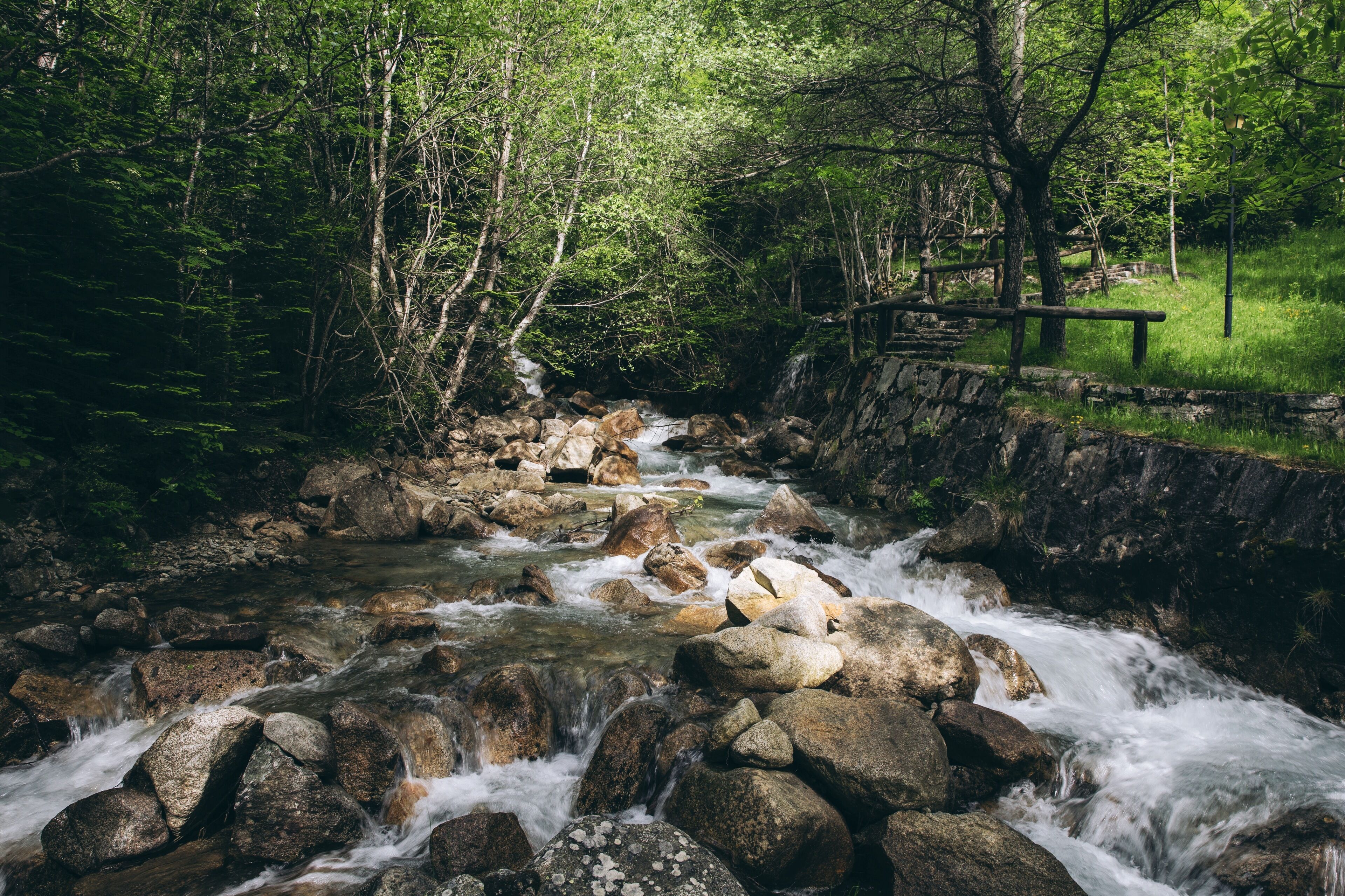 Un arroyo pedregoso que serpentea por un área boscosa, con un muro de piedra y un puente de madera.