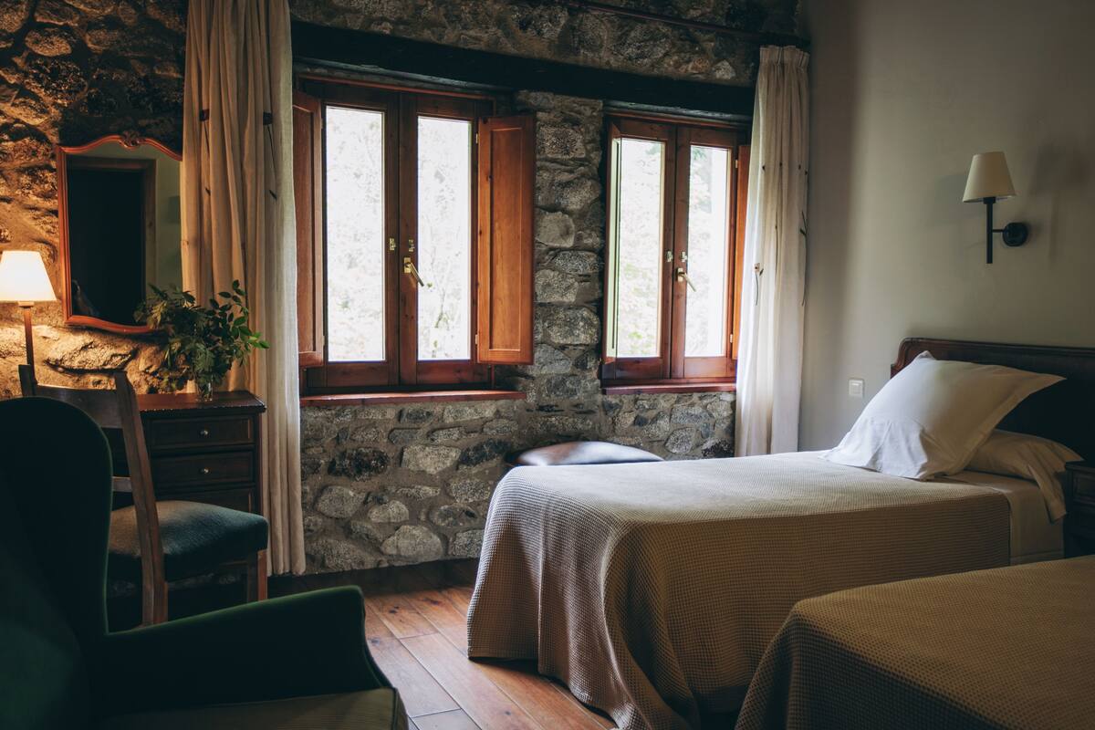 Una habitación con paredes de piedra, camas de madera, un escritorio con lámpara y una silla.