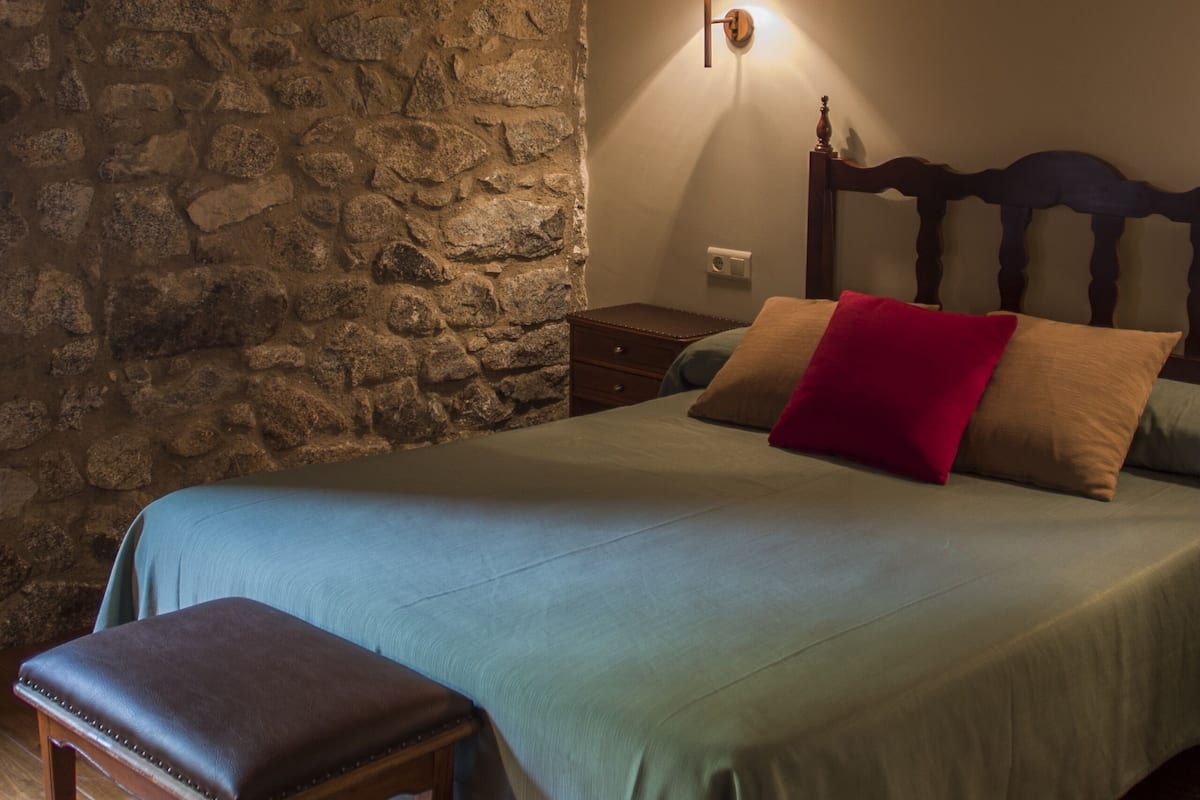 Un dormitorio con pared de piedra, estructura de cama de madera, mesita de noche, lámpara y un banco.