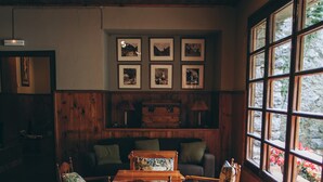 Una habitación acogedora con pisos y techo de madera, un sofá, una mesa y sillas. Se observan cuadros enmarcados en la pared y una ventana con vistas a árboles.