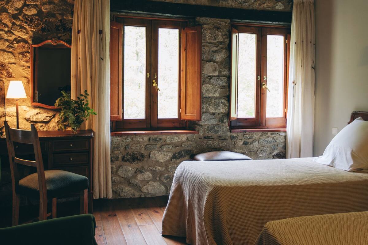 Una habitación rústica con paredes de piedra, vigas de madera, una cama, un escritorio y una silla.