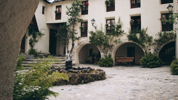Un patio con una fuente, escalones de piedra y un banco. Se ven puertas y ventanas de arco con jardineras.