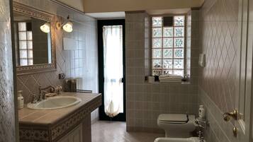 Un baño con dos lavamanos, un inodoro y un bidé. Hay una ventana con vidrios decorativos y un perchero con toallas.