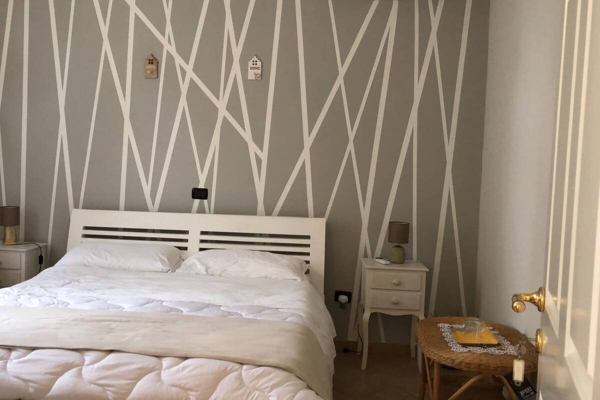 Una cama bien tendida en una habitación con un papel tapiz de diseño geométrico, una mesita de noche con una lámpara y una silla de mimbre.